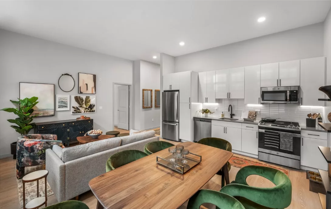 Open-plan kitchen and living area with green velvet chairs, a wood dining table, and modern white cabinetry with stainless steel appliances.