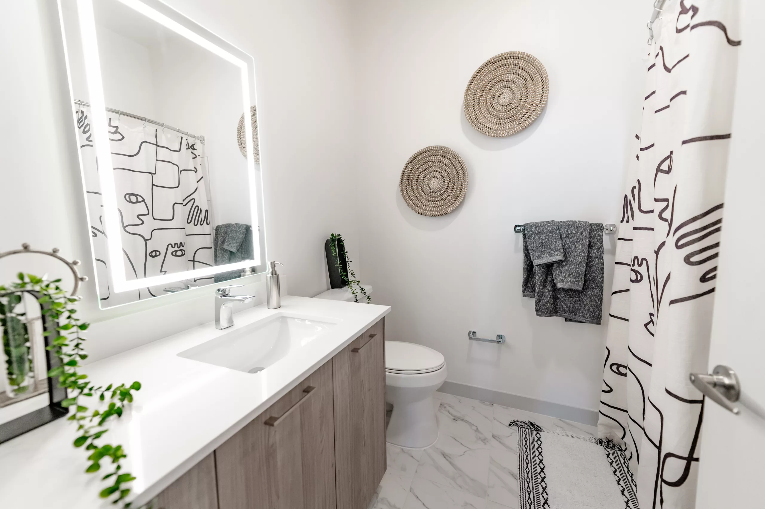 Bathroom with backlit mirror, abstract shower curtain, and woven wall decor