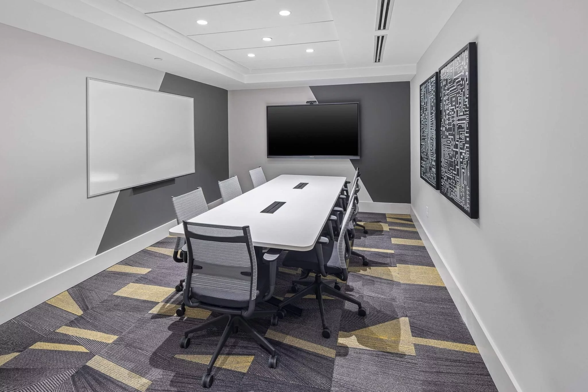 Modern conference room with a long white table, office chairs, wall-mounted screen, and patterned carpet.