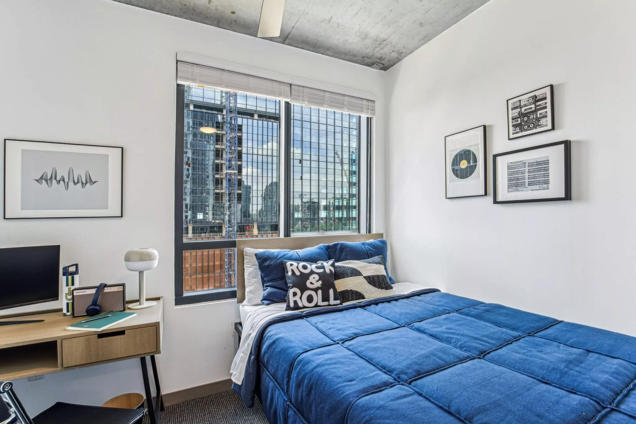 Bedroom with large window, blue bedding, wall art, desk, and a view of city buildings under a concrete ceiling.