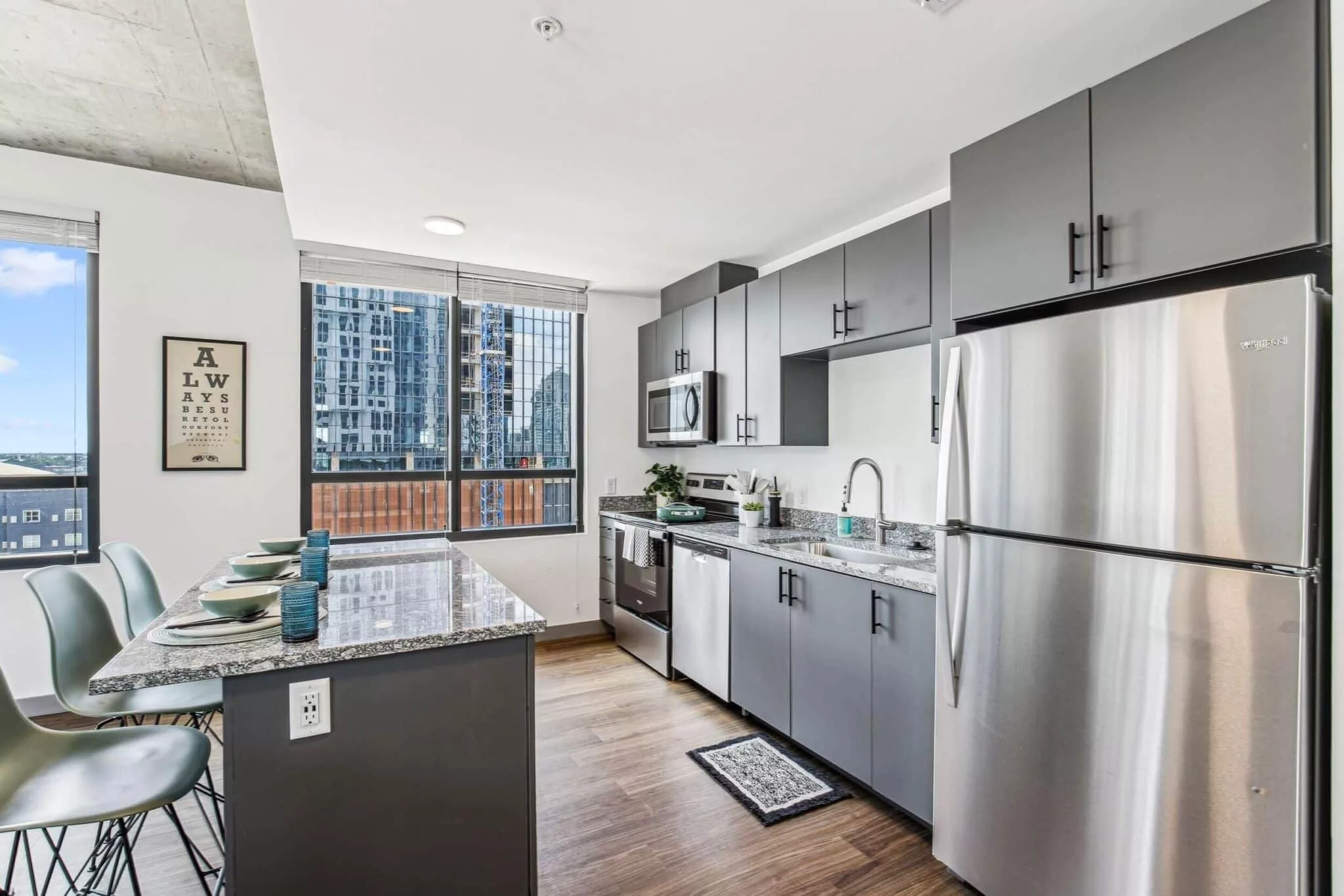 Modern kitchen with stainless steel appliances, grey cabinetry, granite countertops, and city views through large windows.