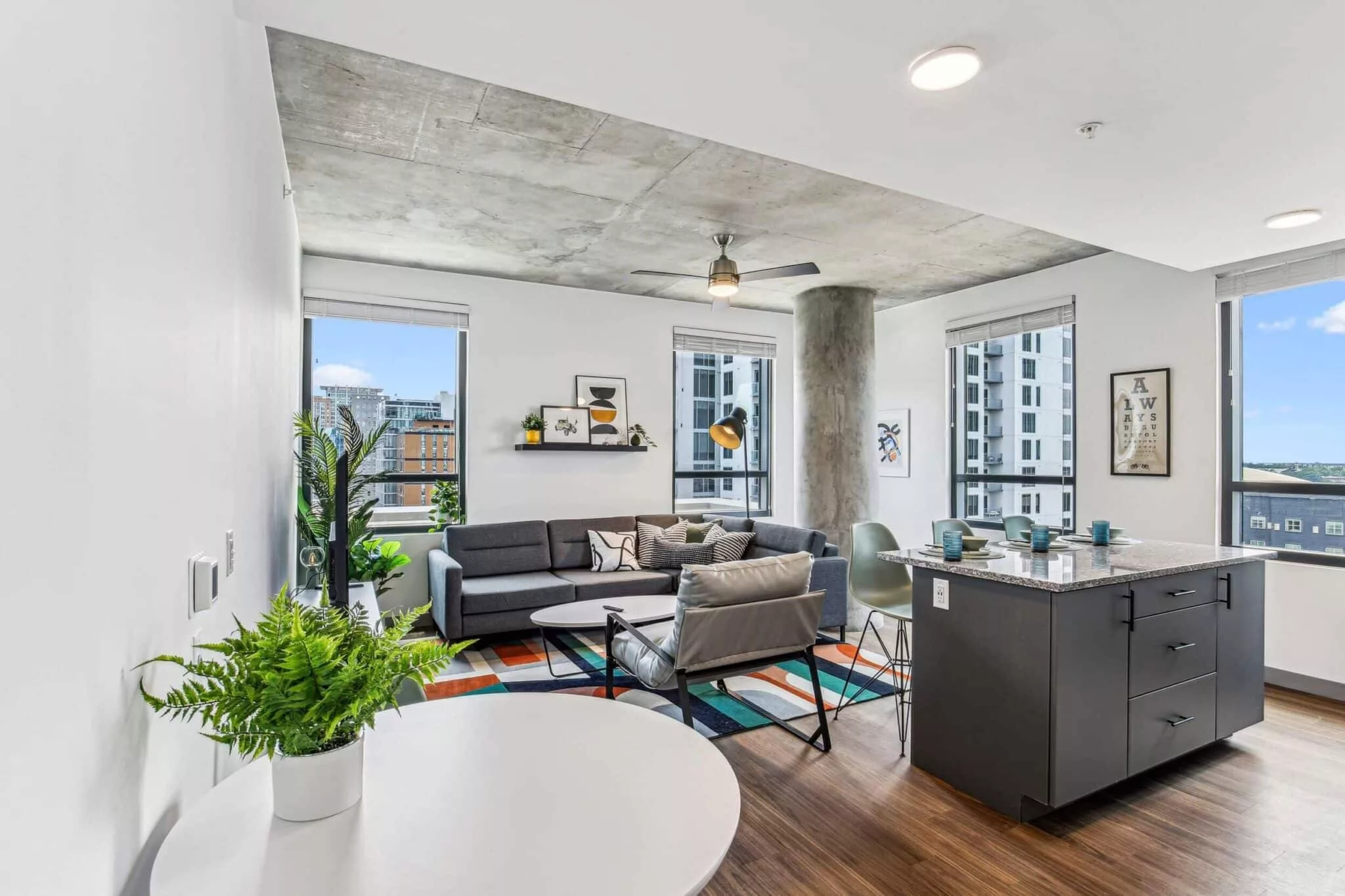 Open-concept living and dining area with grey sectional, modern furniture, and floor-to-ceiling windows with urban views.