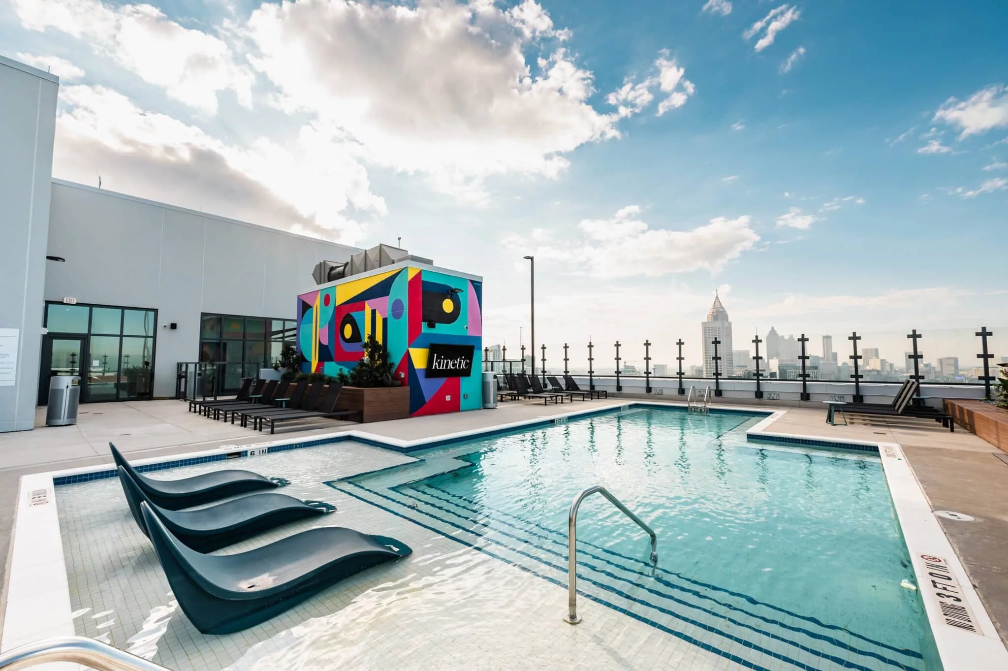 Rooftop swimming pool with in-water lounge chairs, city skyline views, and a colorful kinetic mural wall.