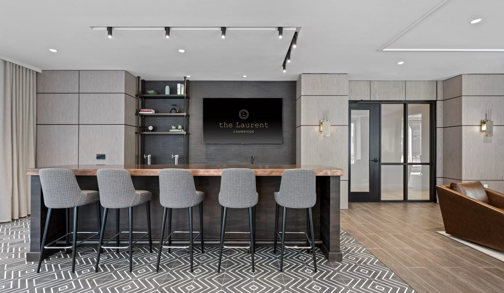 Lounge bar area with upholstered high stools, a wooden countertop, and a large display reading "The Laurent Cambridge."