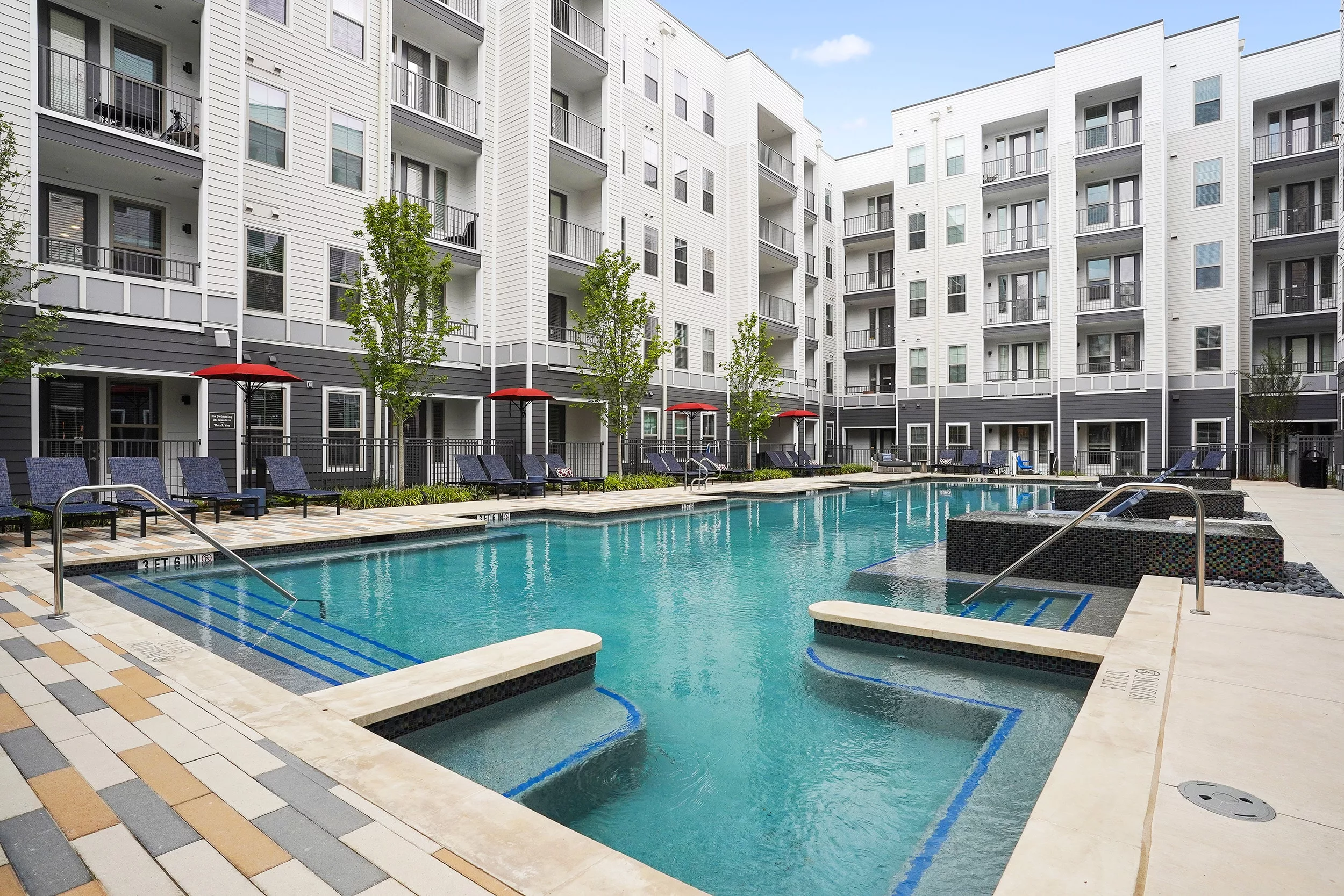 Modern apartment courtyard with large swimming pool, lounge chairs, red umbrellas, and white and gray multi-story buildings.