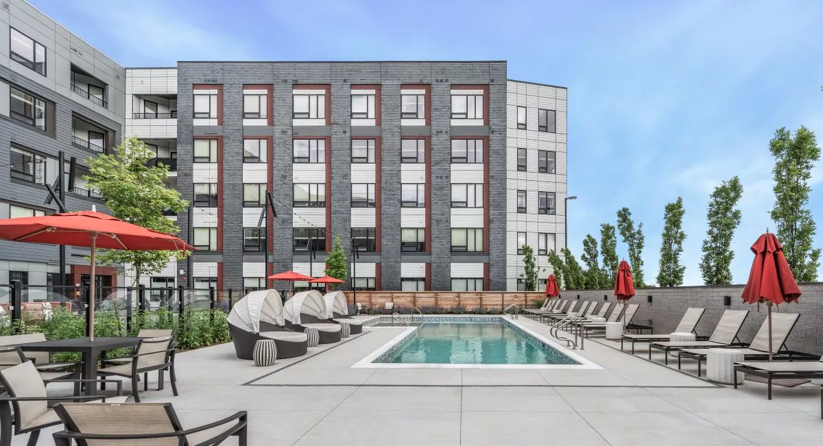 An outdoor pool surrounded by lounge chairs with red umbrellas outside of a gray and red apartment building.
