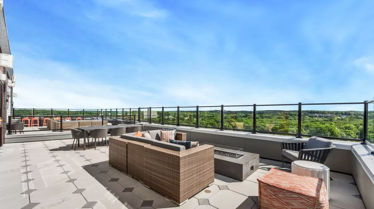 A rooftop patio with various seating around a firepit, a long table with chairs, and views of treetops under a blue sky.