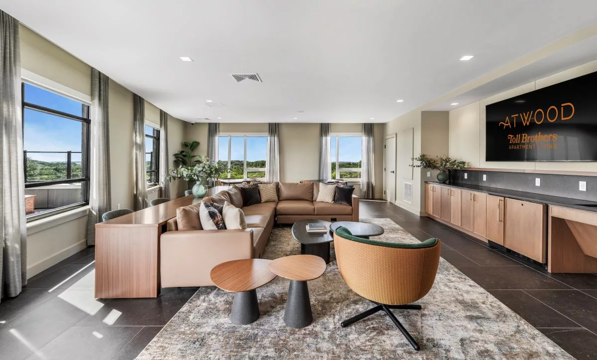 A lounge with large windows, beige walls, a large brown sectional soda, and small wooden tables.