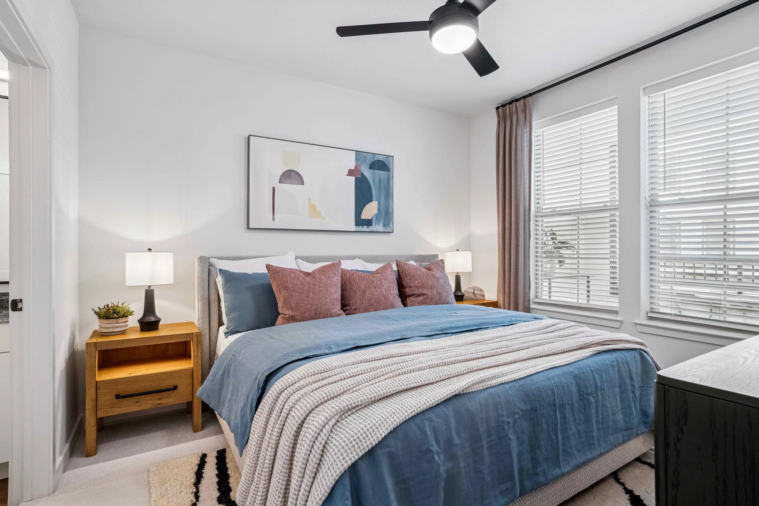 Model bedroom with blue bedding, nightstands, lamps, a ceiling fan, and windows with blinds.