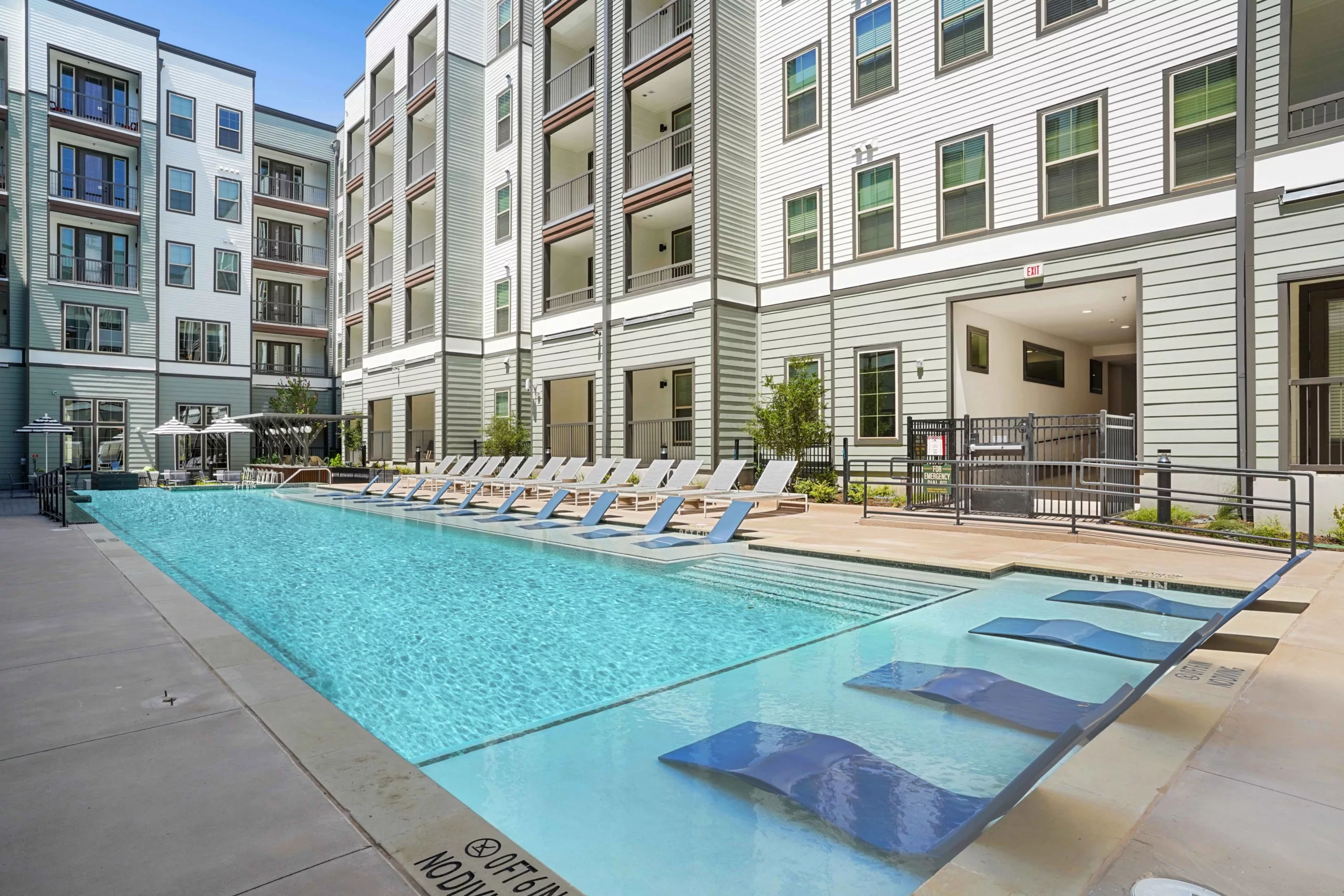 Resort-style pool with in-water loungers and deck chairs, adjacent to a multi-story building.