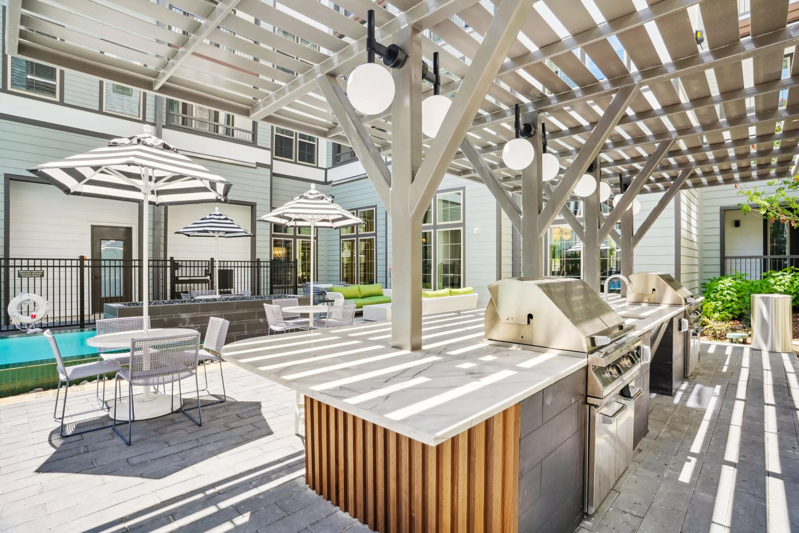 Outdoor kitchen with stainless steel gas grills under a pergola with globe lights. Patio seating nearby.