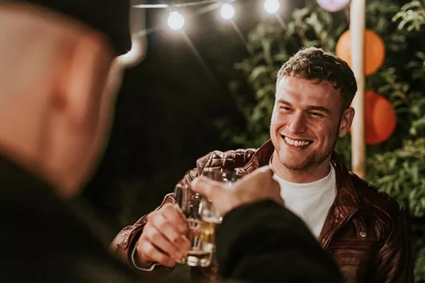 Two people clinking beer glasses at an outdoor party with string lights and decorations.