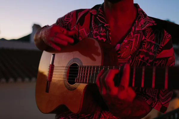Person playing an acoustic guitar at sunset, wearing a patterned shirt.